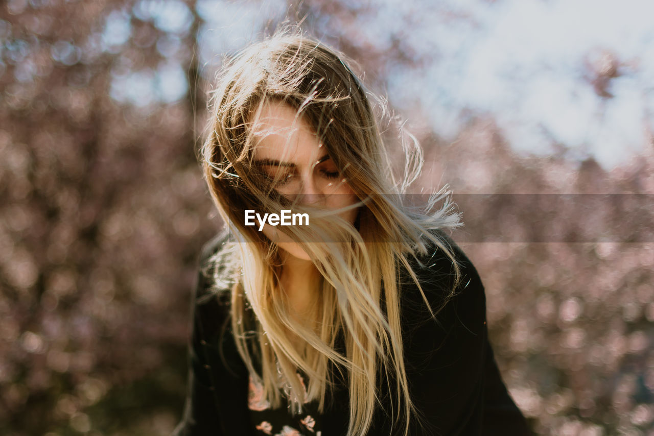 Beautiful young woman looking down with tousled hair standing against trees