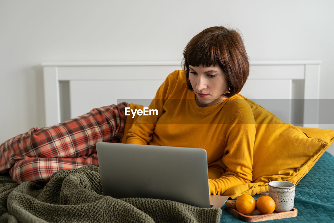 Smiling woman using laptop while sitting on bed at home