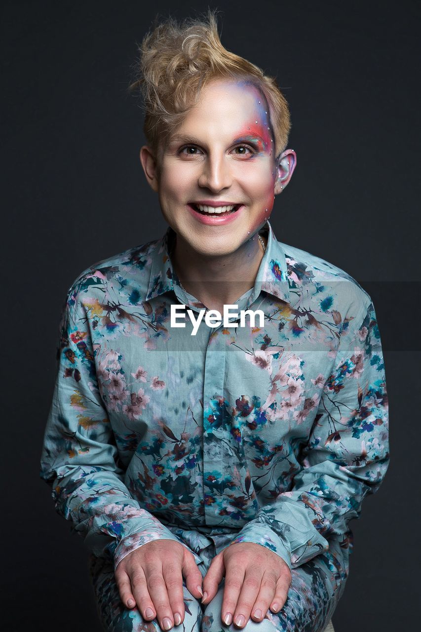 Portrait of smiling young man with make-up in floral clothing against black background