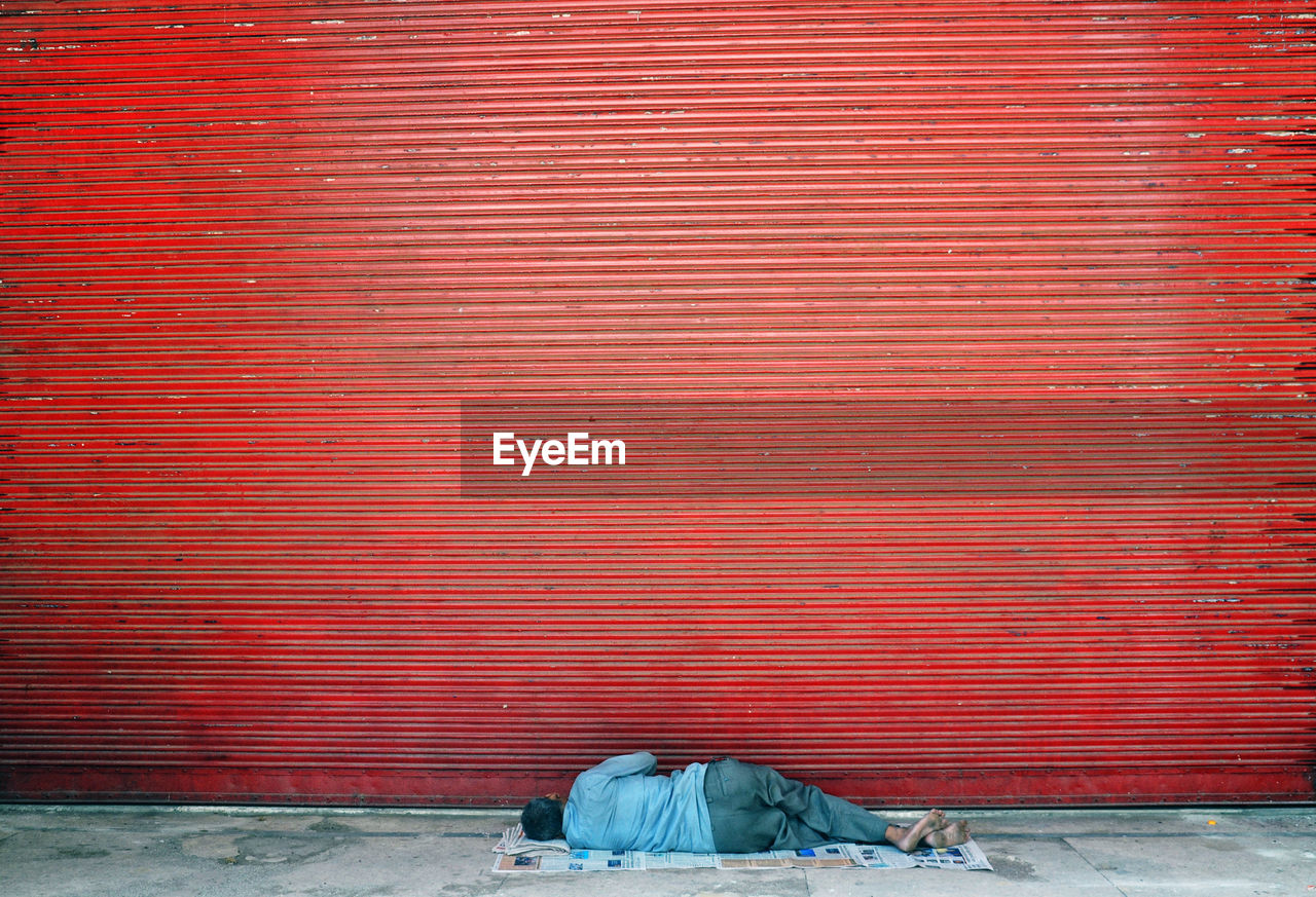 Rear view of homeless man sleeping on footpath by closed red shutter