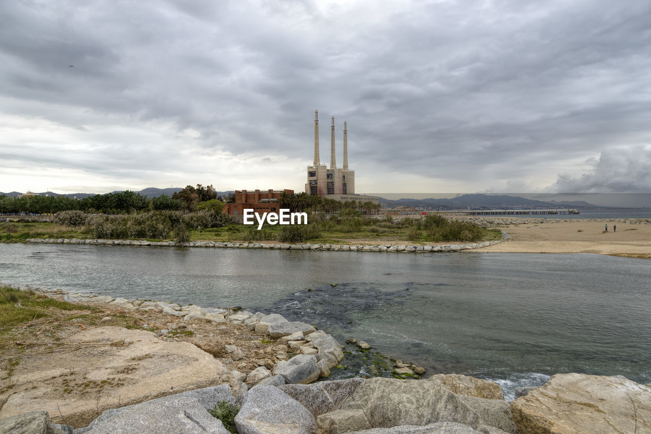 VIEW OF RIVER BY FACTORY AGAINST CLOUDY SKY