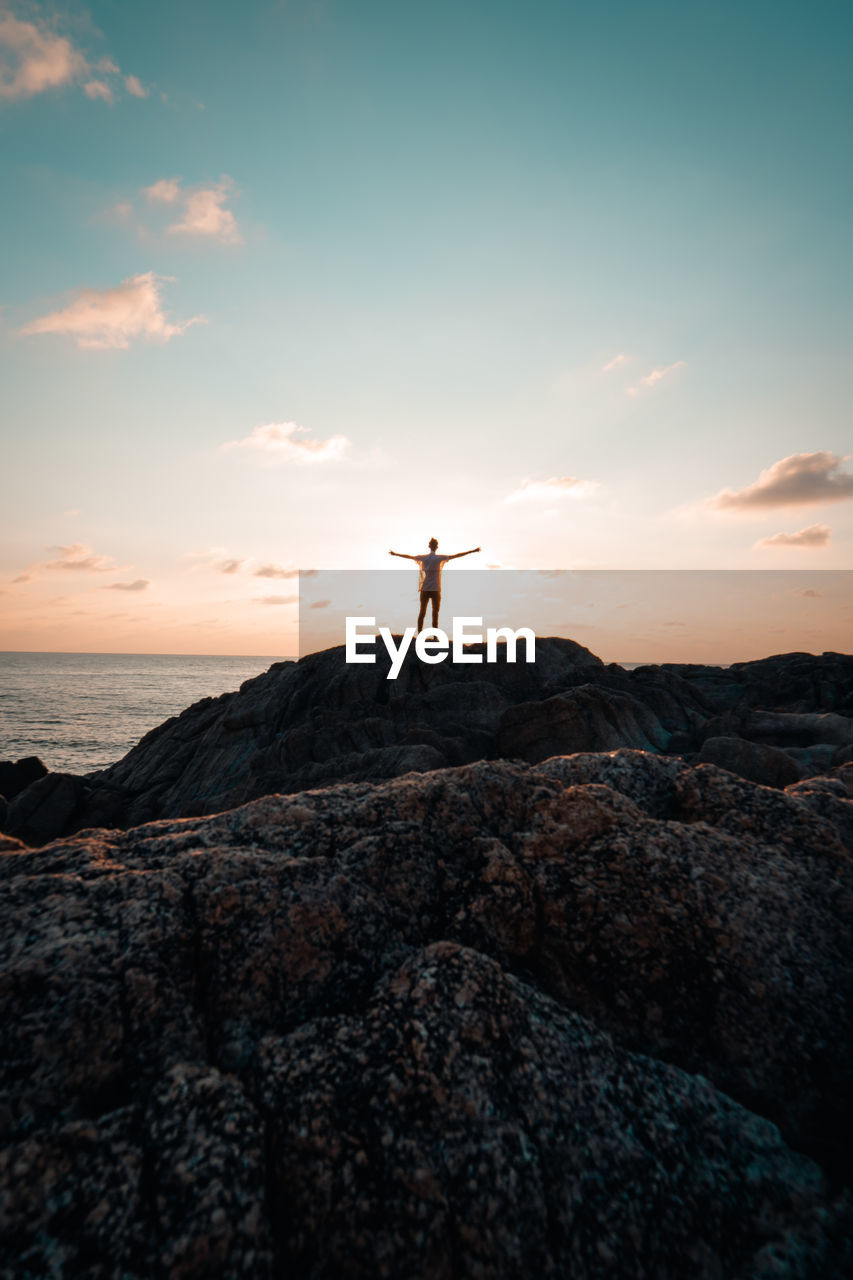 Distant view of person standing on rocky shore against cloudy sky during sunset