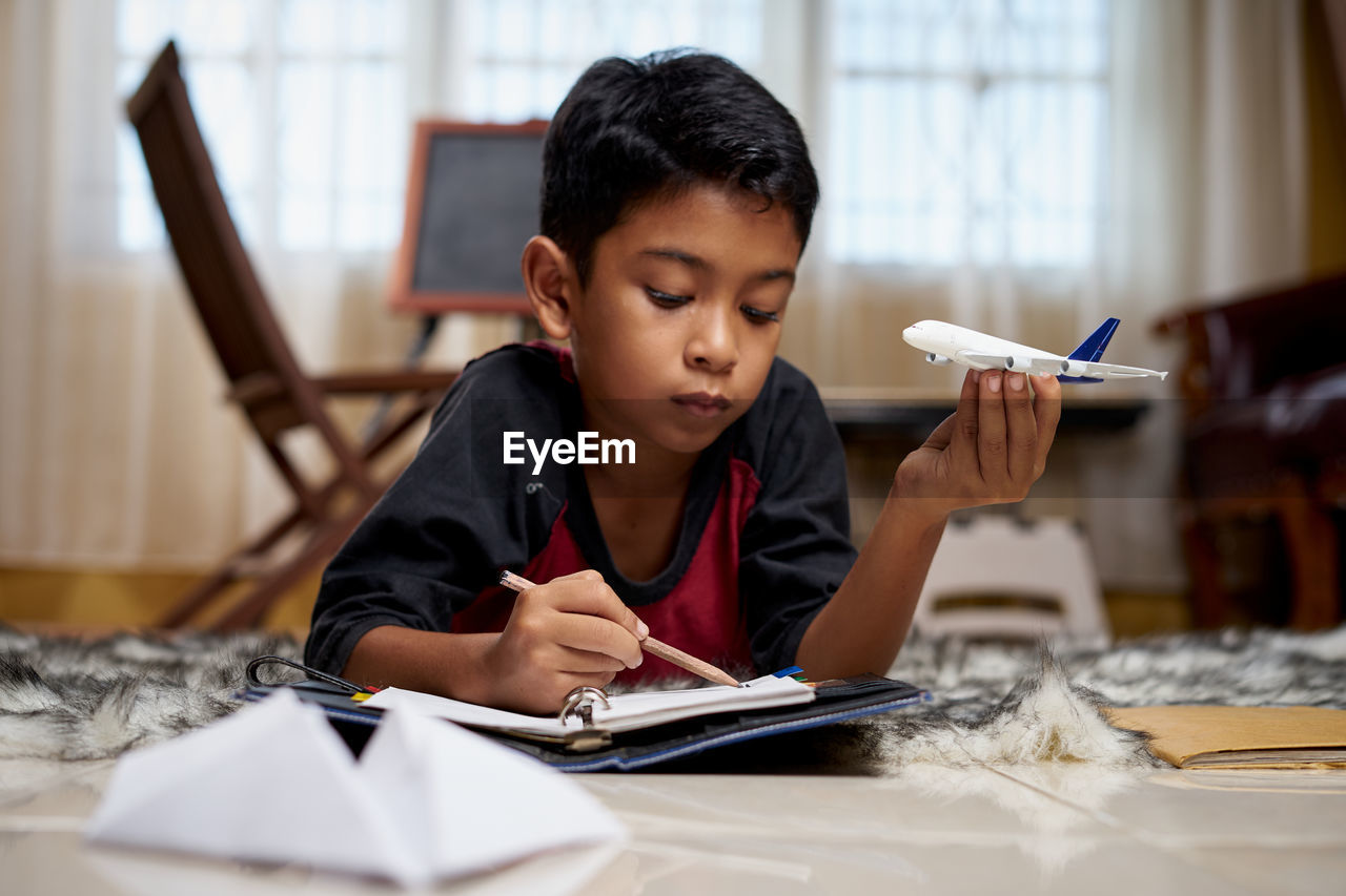 Contemplating boy writing while holding model airplane on bed