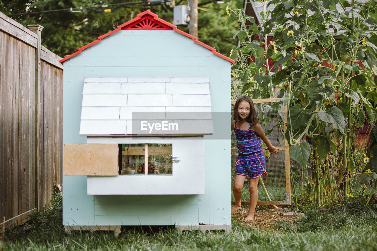 A proud little girls stands at the open door of chicken coop in garden