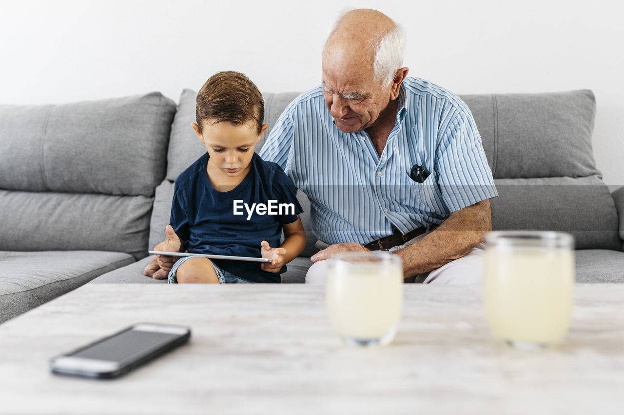 Grandfather and grandson sitting together on the couch at home looking at digital tablet