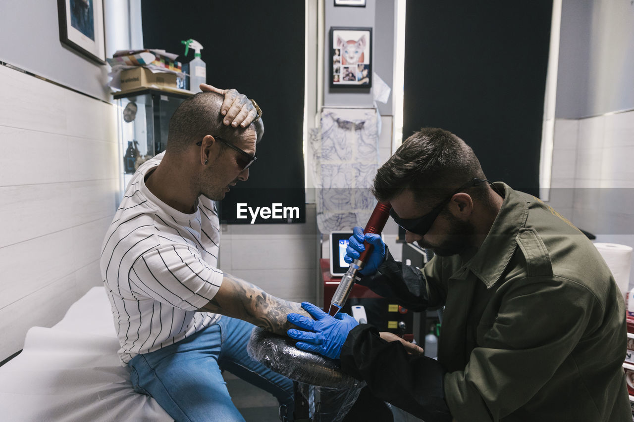 Side view of focused male making laser tattoo on arm of tired shaved client in tattoo salon