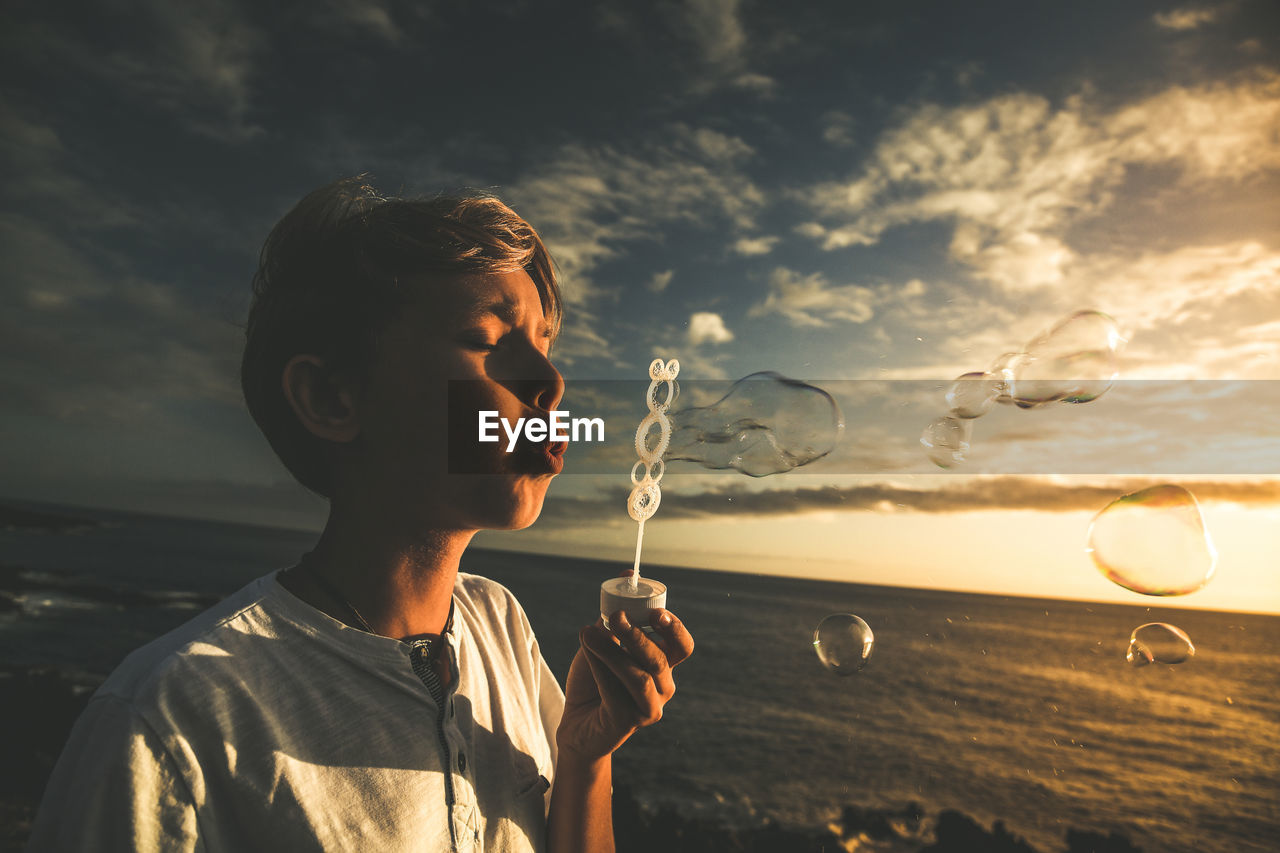 Close-up of cute boy blowing bubbles against sky during sunset