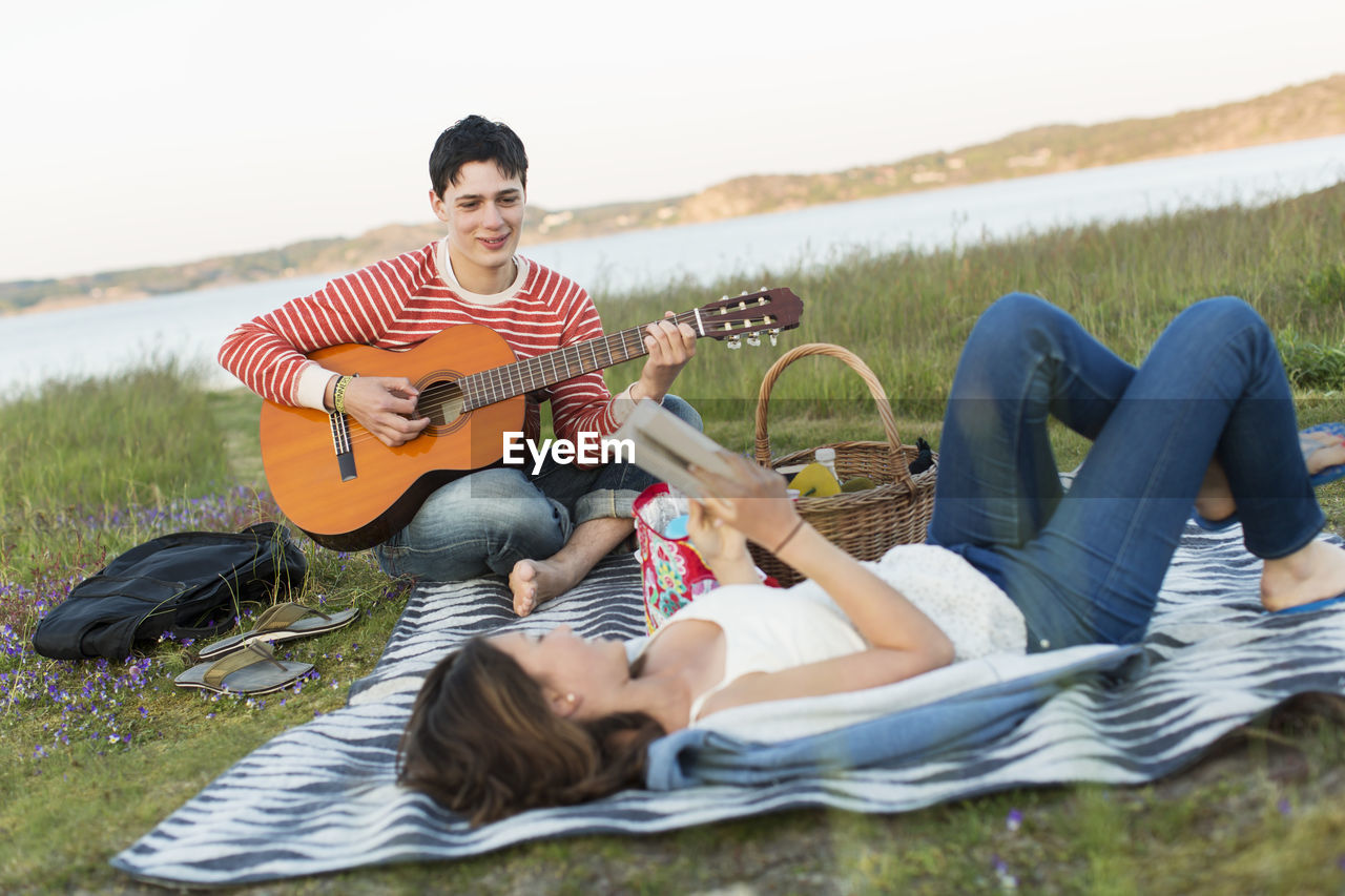 Teenage girl reading book with boyfriend playing guitar during picnic