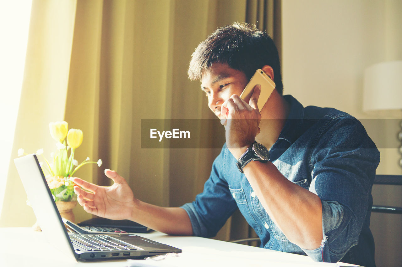 Young businessman talking on phone while looking at laptop in office