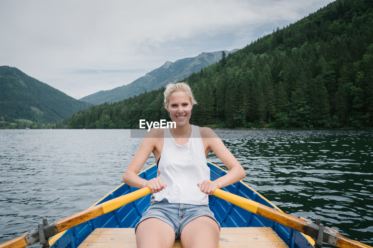 PORTRAIT OF SMILING YOUNG WOMAN SITTING ON LAKE