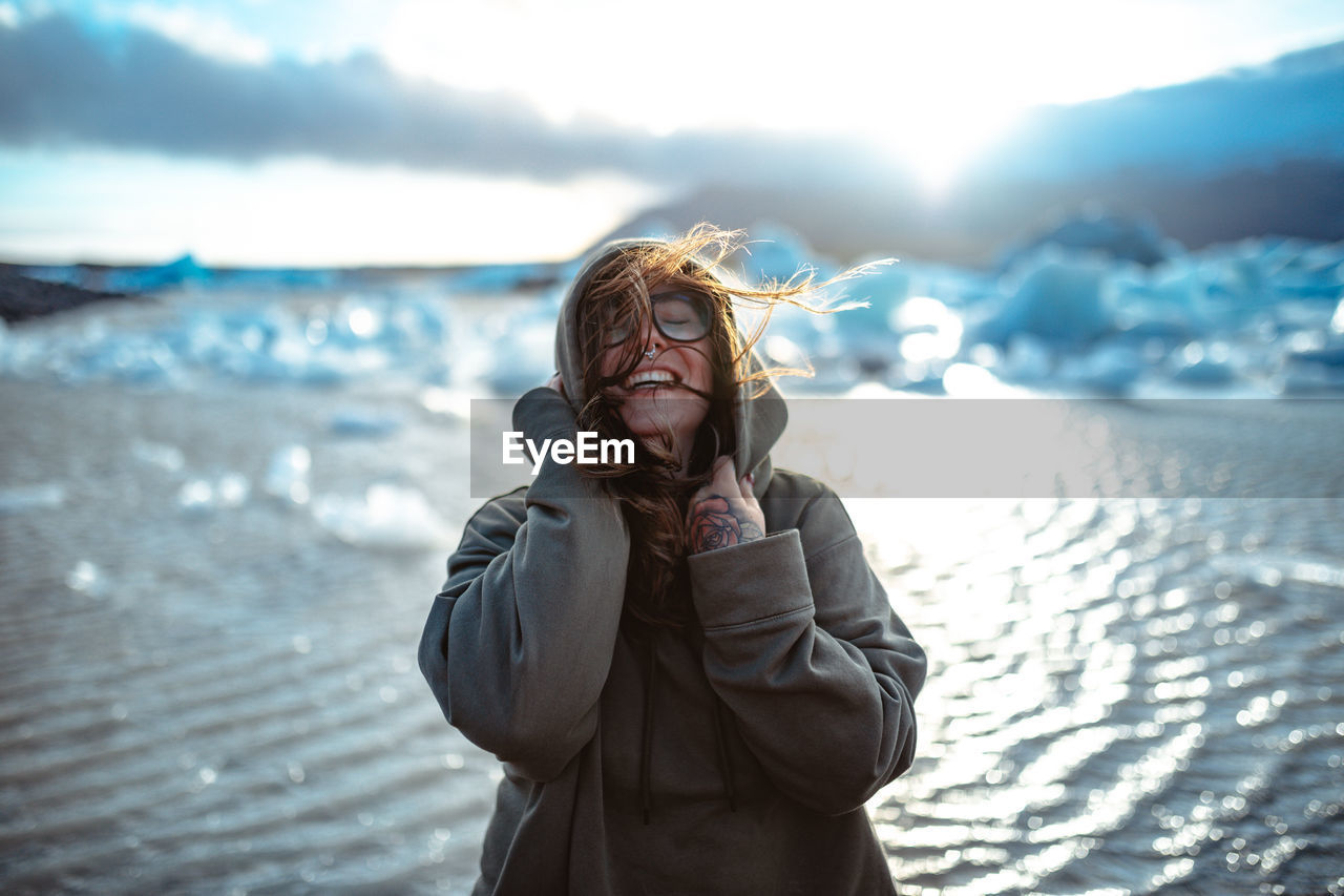 Young laughing tourist in eyeglasses with piercing and windy hair near water in sunny day on blurred background