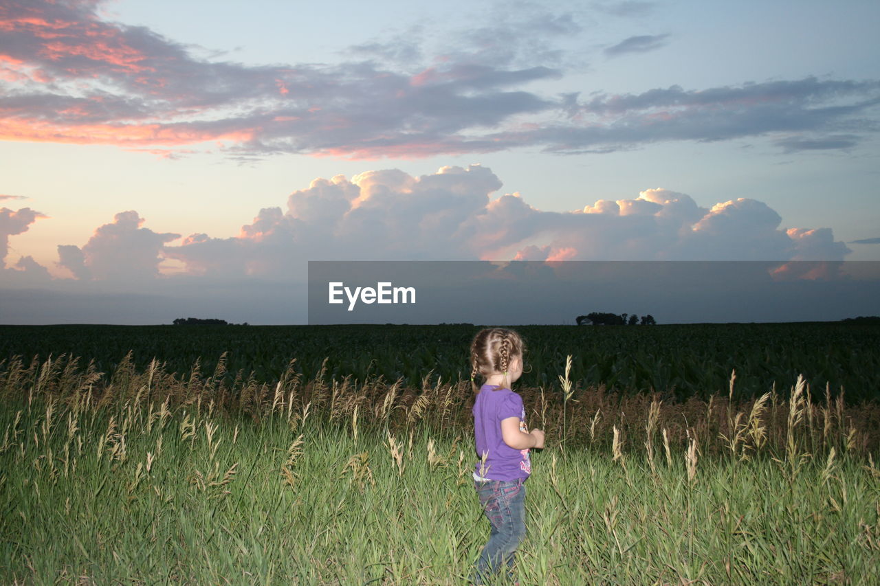 Side view of girl standing on grassy field against sky during sunset