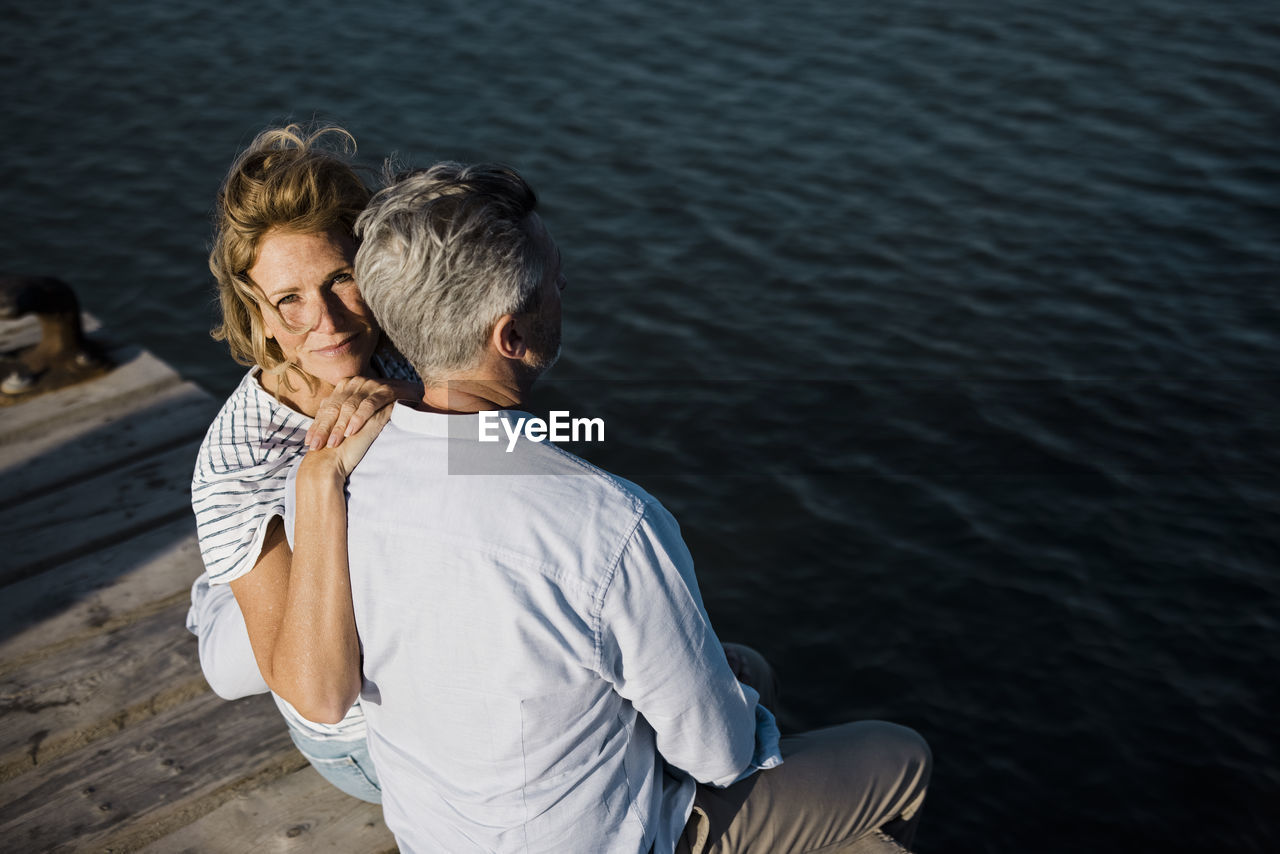 Smiling mature woman sitting with hand on man's shoulder at jetty
