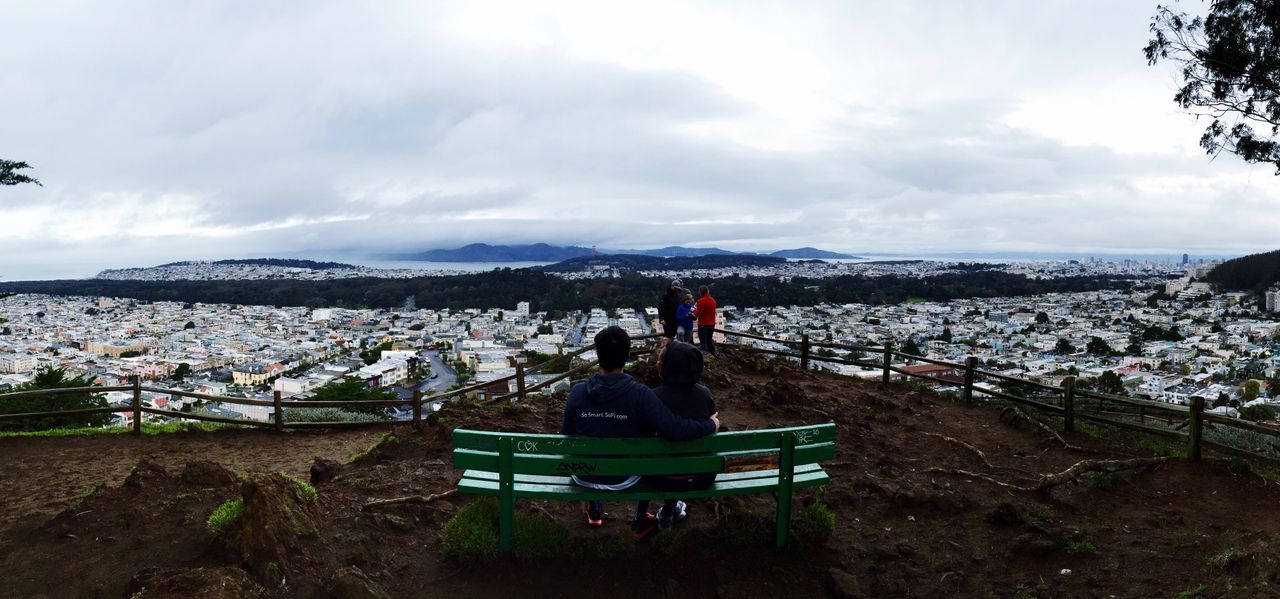 REAR VIEW OF MAN SITTING ON LANDSCAPE AGAINST CLOUDY SKY