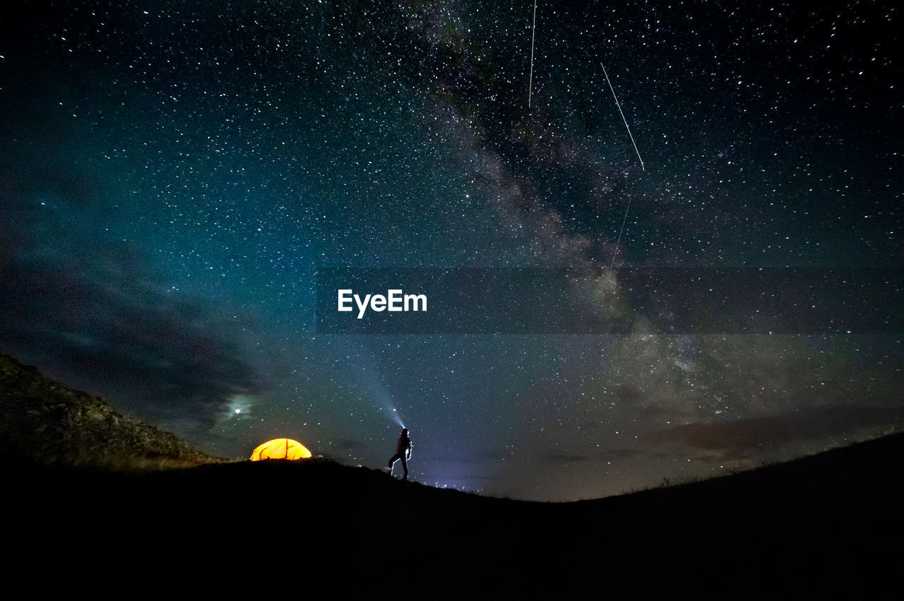 Star fall at the night sky and a silhouette of a man and an orange tent on the horizon