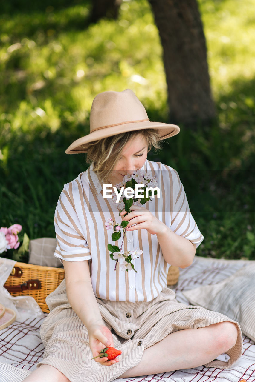 sitting, hat, plant, adult, women, nature, sun hat, leisure activity, relaxation, clothing, summer, spring, smiling, one person, food and drink, lifestyles, fashion accessory, happiness, casual clothing, emotion, person, full length, straw hat, day, grass, outdoors, female, food, picnic, tree, flower, holding, fedora, front view, young adult, sunlight, flowering plant, enjoyment, seat, child, park, three quarter length, wellbeing, childhood, positive emotion, bench, park - man made space, holiday, looking