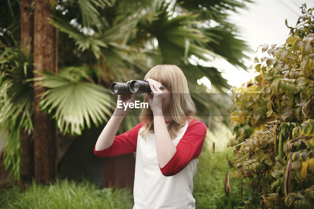 Teenager looking through binoculars while standing by plants at park