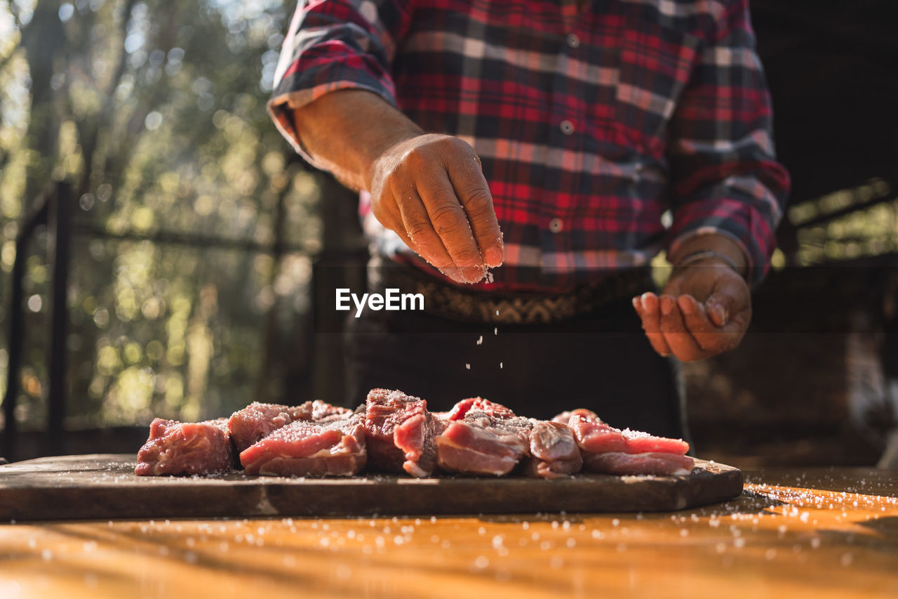 Crop unrecognizable man in checkered shirt putting salt on pieces of meat on wooden cutting board while preparing food in woods