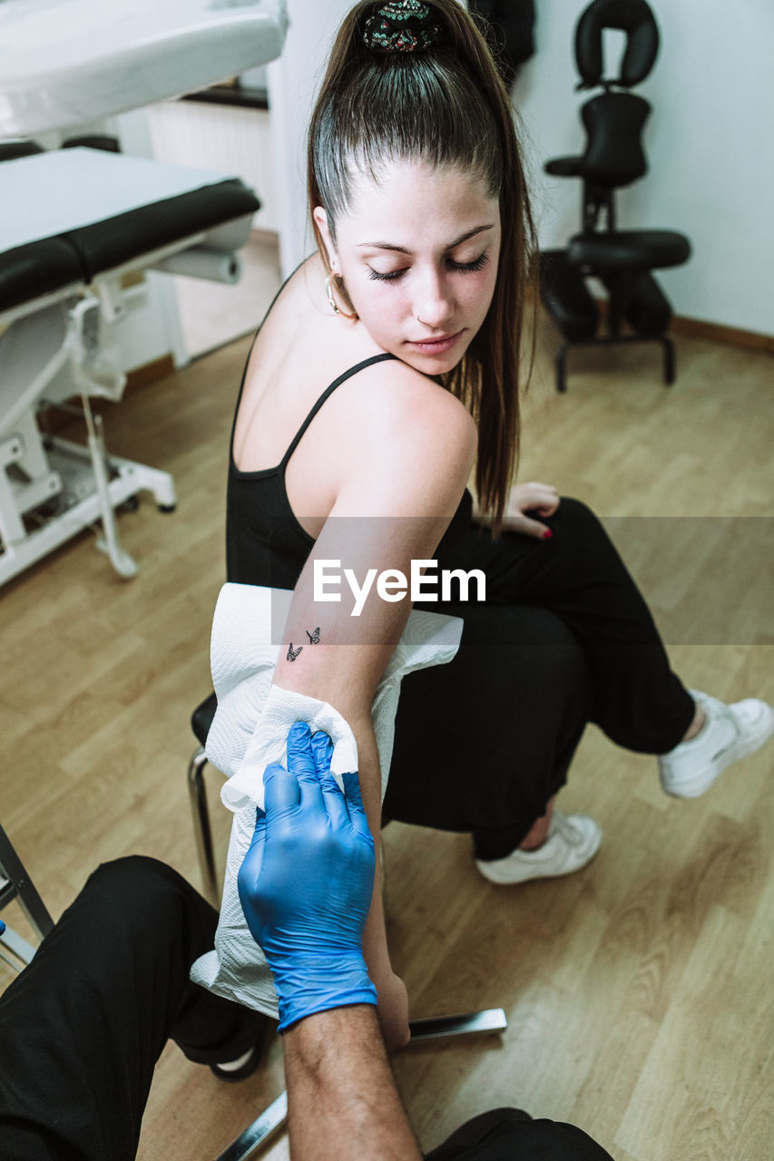 Back view crop anonymous male tattooist in latex gloves wiping freshly made tattoo on arm of female client with napkin while working in salon