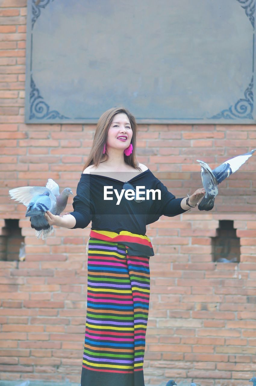 Portrait of woman with birds standing against brick wall