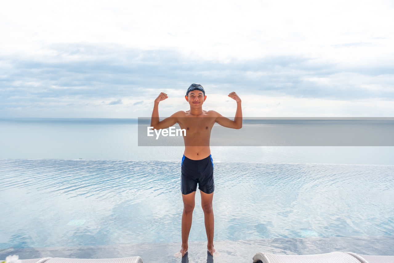 FULL LENGTH OF SHIRTLESS MAN STANDING ON BEACH AGAINST SKY