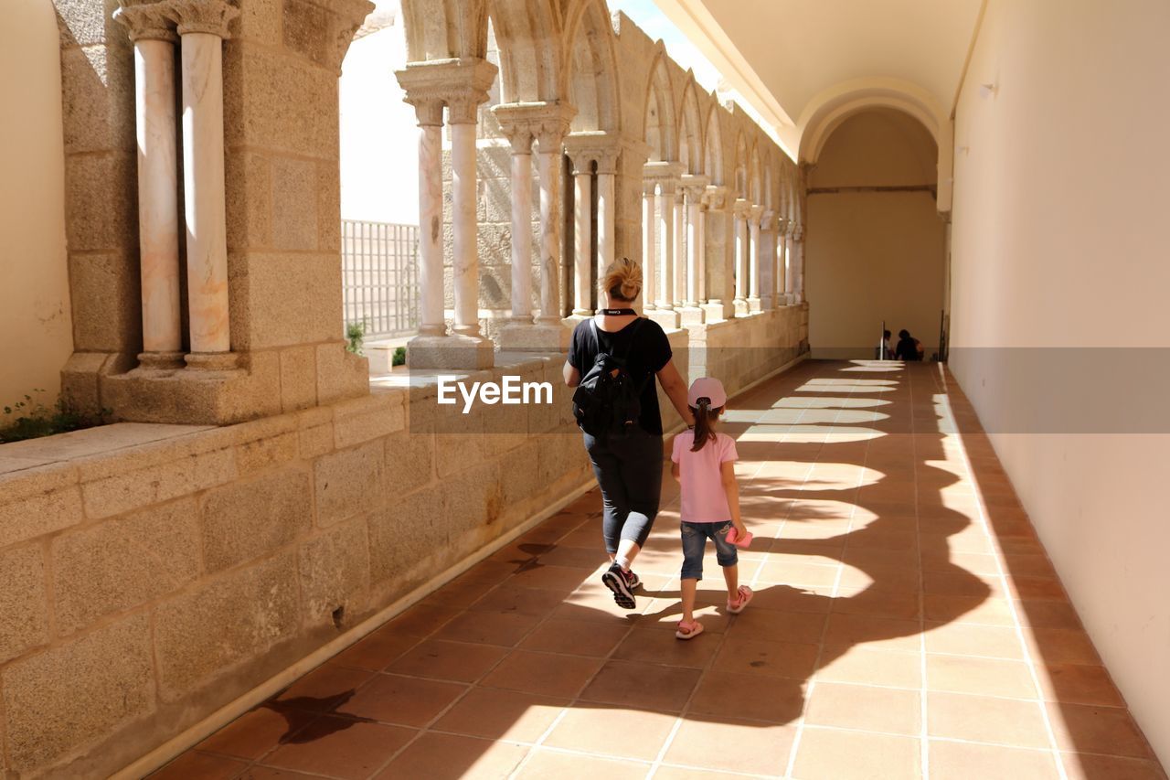 Rear view full length of mother with daughter walking in historic building passage