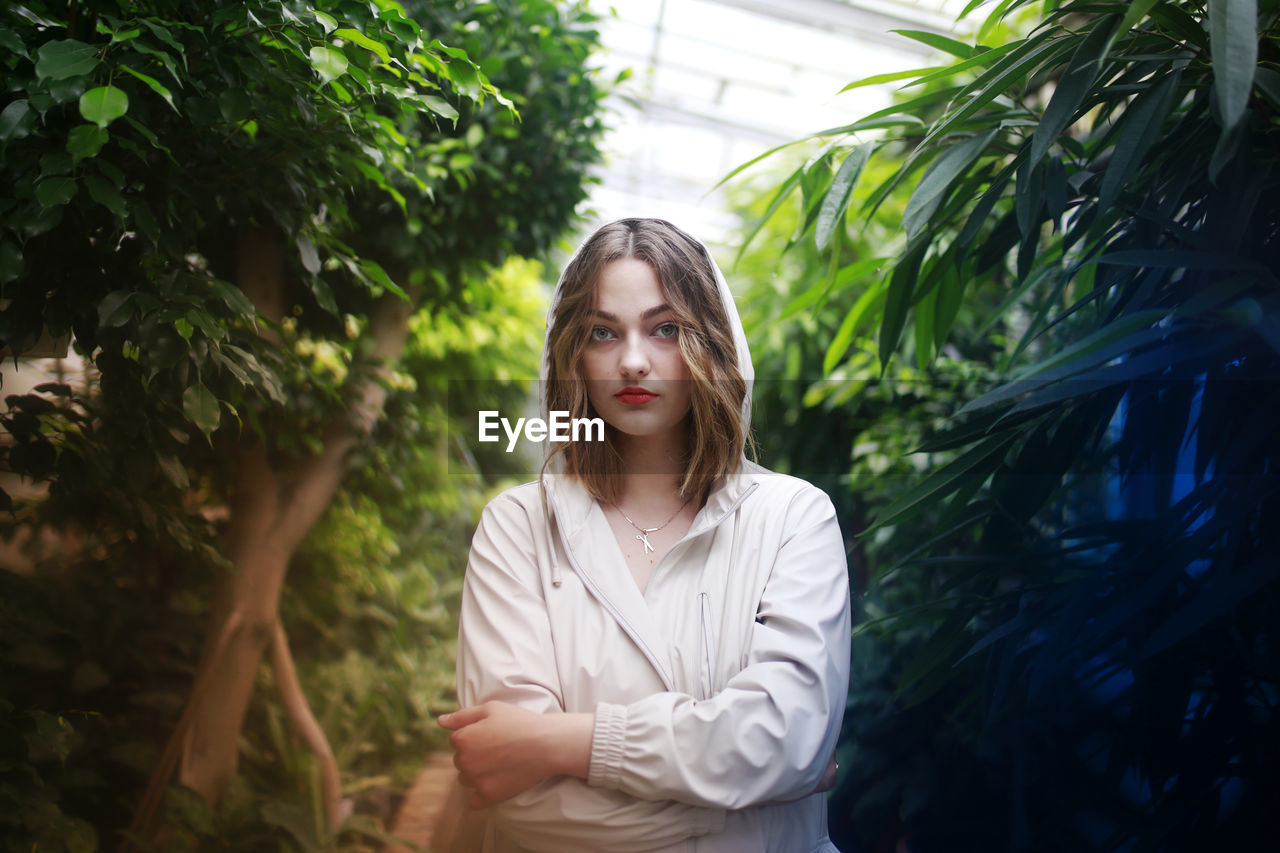 PORTRAIT OF YOUNG WOMAN STANDING BY PLANTS