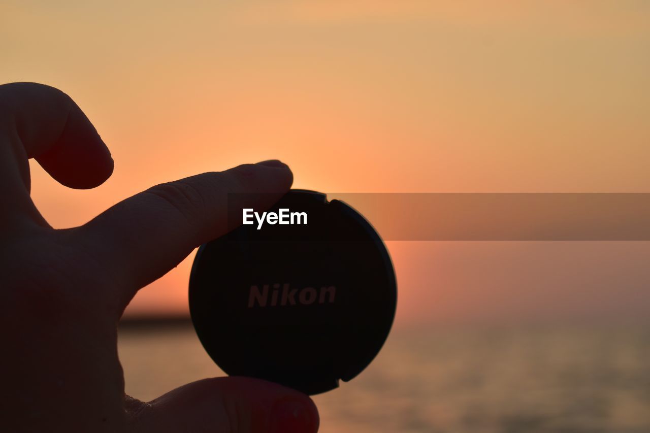 Close-up of hand holding plastic container against sky during sunset