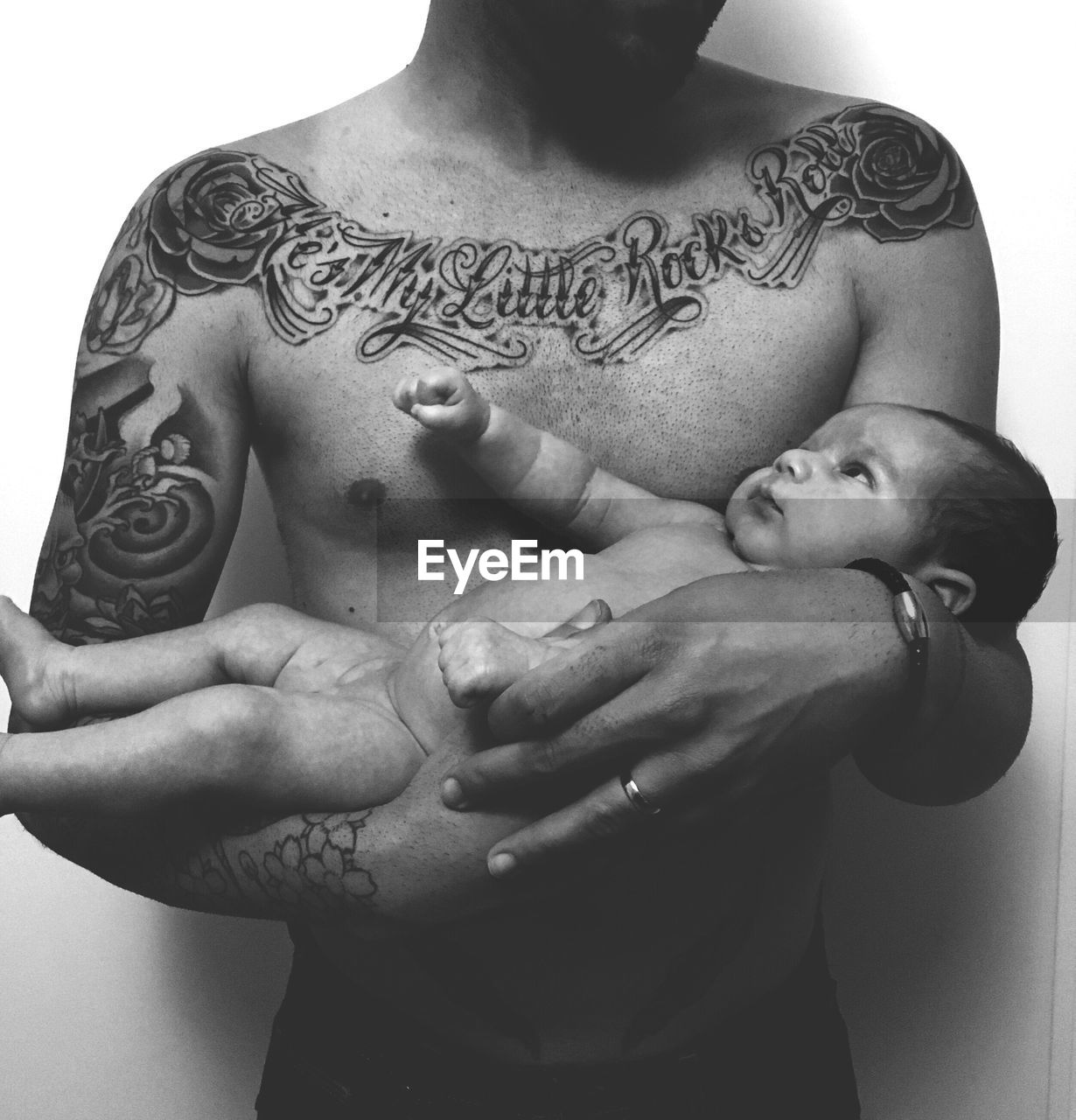 Mid section of shirtless tattooed man holding baby against wall