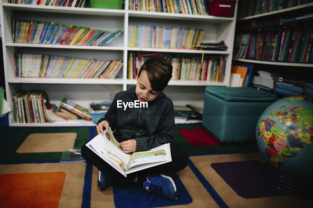 Boy studying while sitting against bookshelves in library