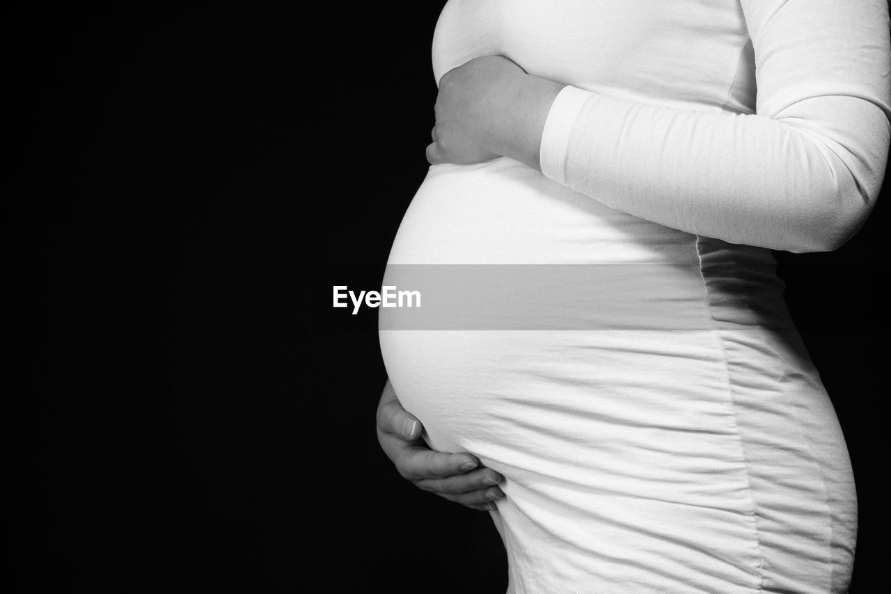Midsection of pregnant woman touching abdomen and standing against black background 