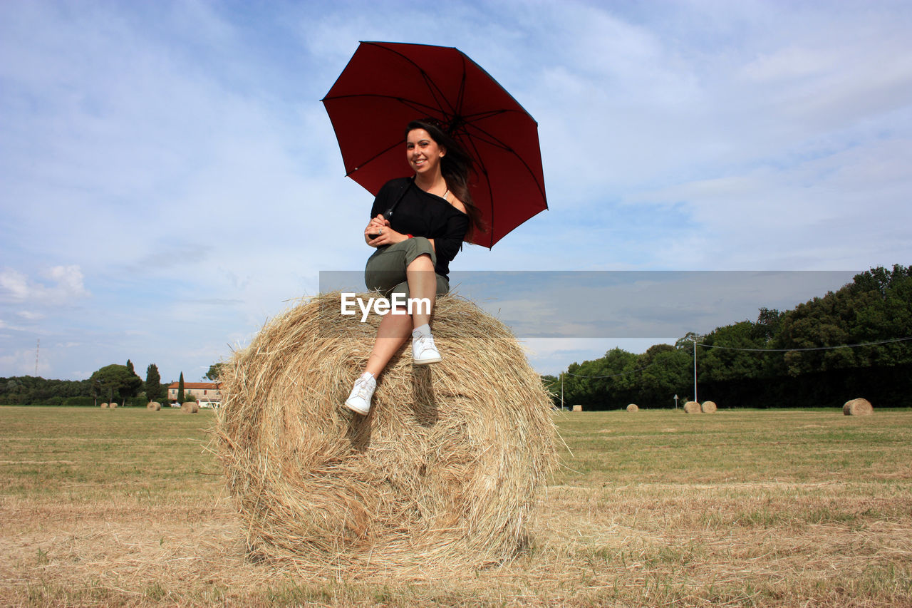 Young woman on a hay ball in a plowed field in tuscany in summer under the umbrella enjoying the sun