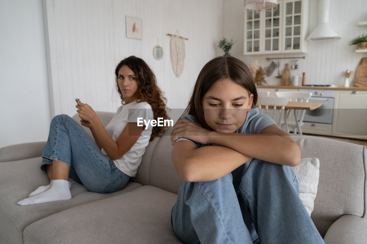 Casual worried woman sits on sofa behind sad teen girl leans on knees looks into distance feels