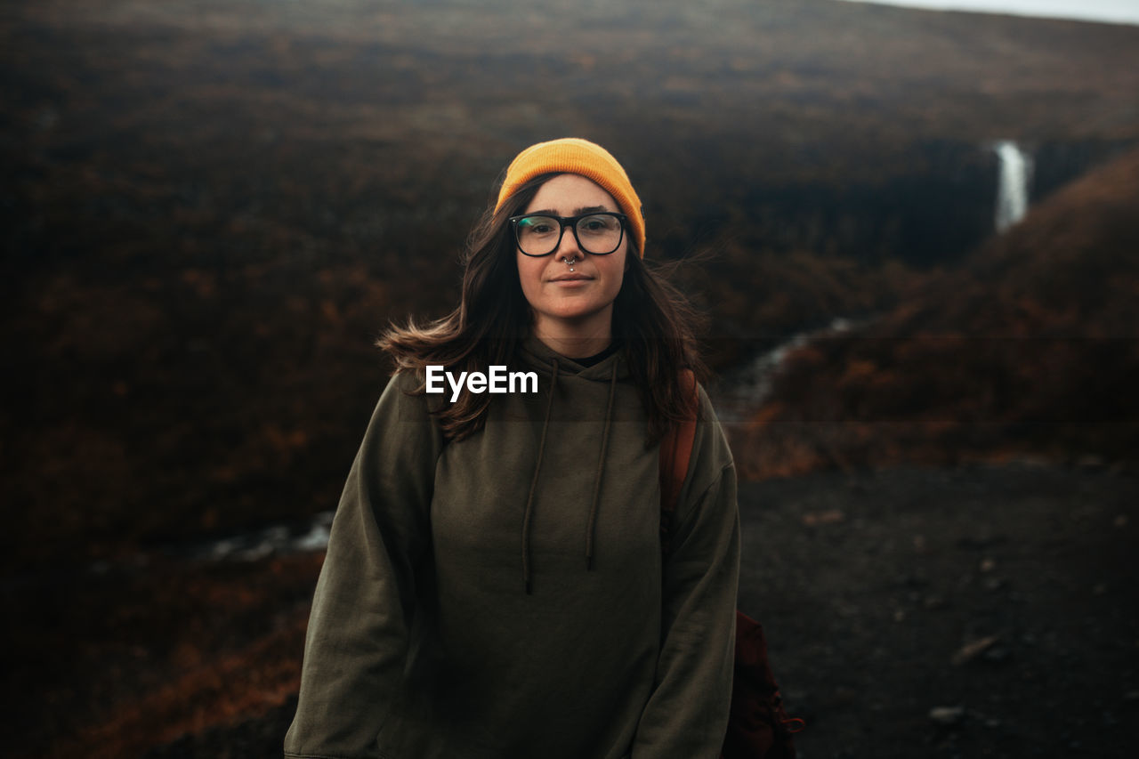 Young tourist in eyeglasses and hat with piercing looking at camera on hill near waterfall and mountain river