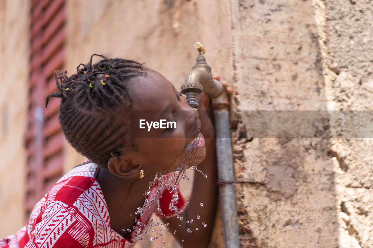 Girl drinking water from faucet against wall