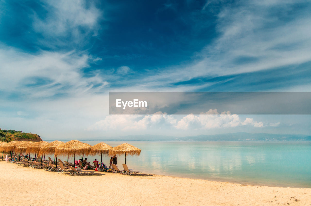 Thatched roof umbrellas and lounge chairs at beach against cloudy sky