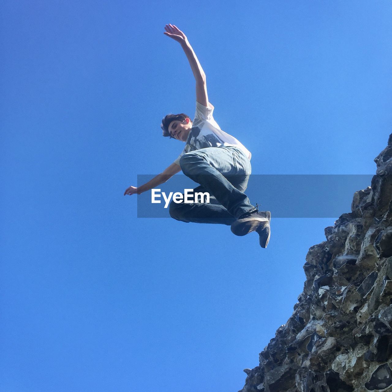 Low angle view of boy jumping from retaining wall against clear blue sky