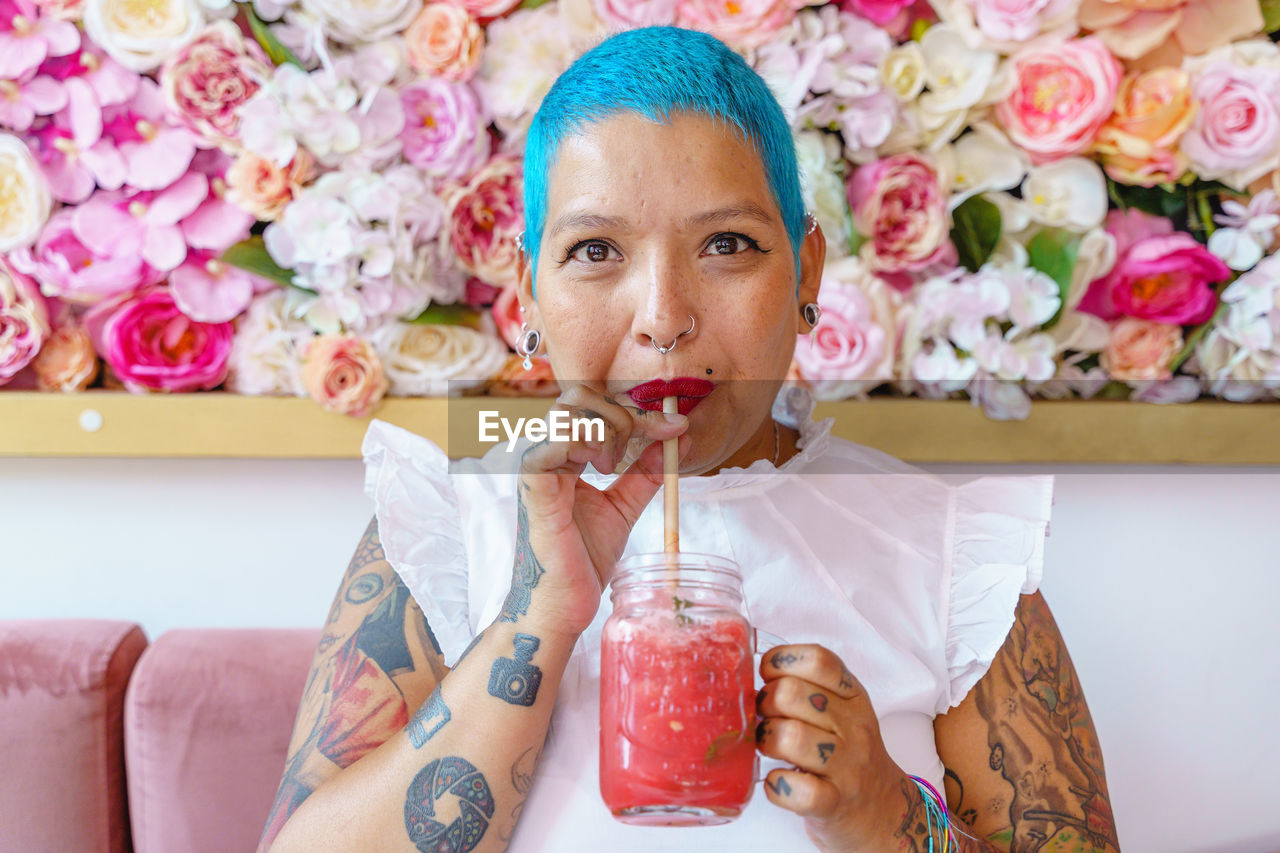 Soft focus of glad female with dyed hair and tattoos sitting in cafe drinking glass jar of fresh nonalcoholic beverage with straw