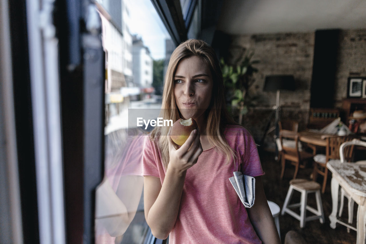 Young woman leaning on window, eating apple