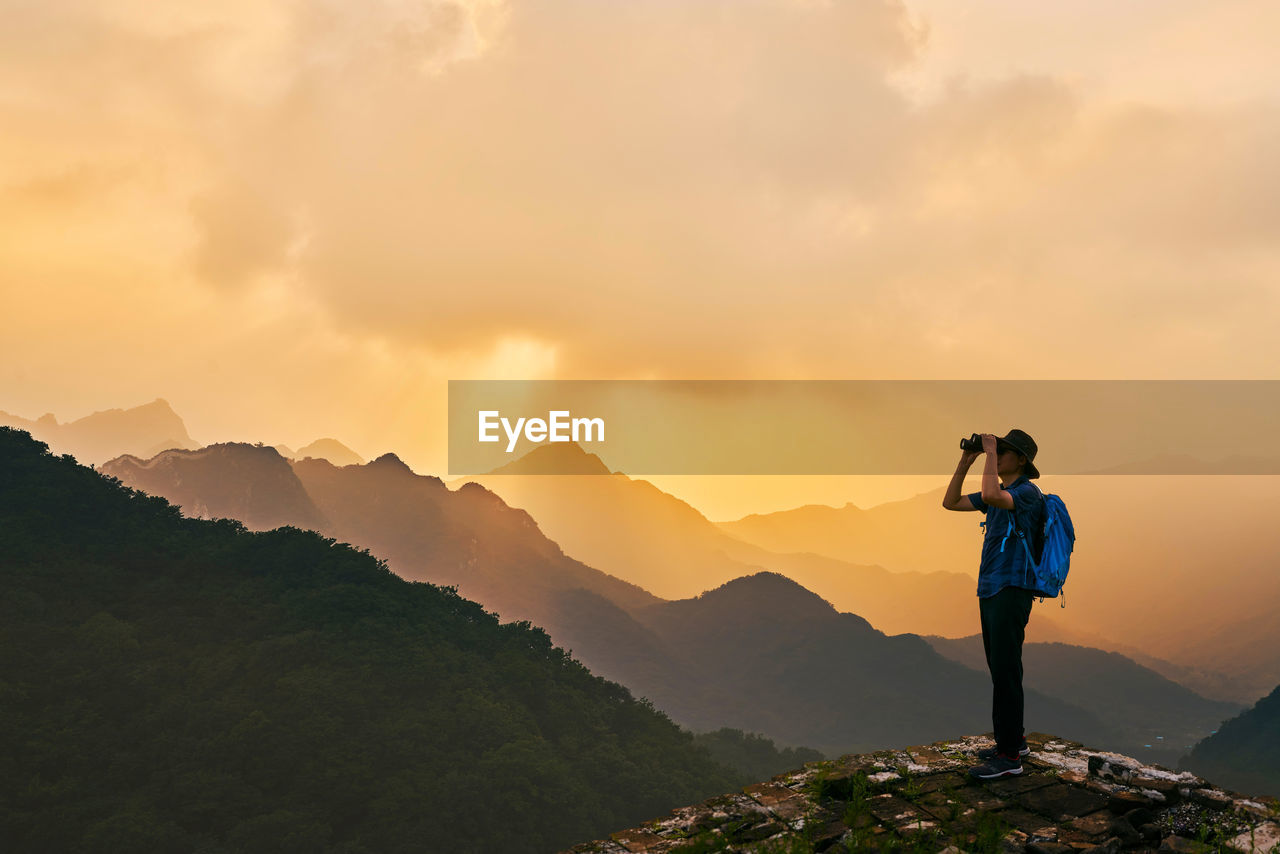Man looking through binoculars while standing on mountain against sky during sunset