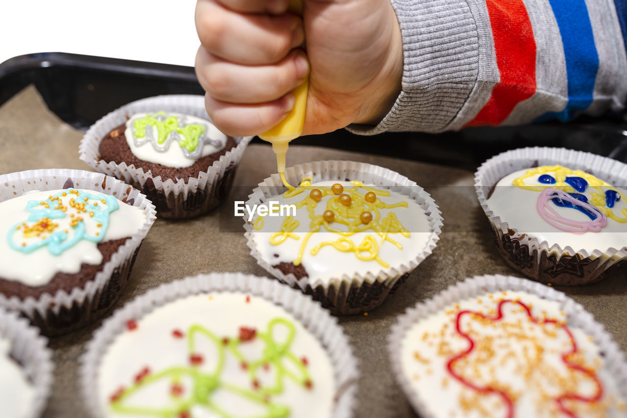 A child squeezes colored frosting from a tube onto chocolate brown cupcakes covered white frosting.