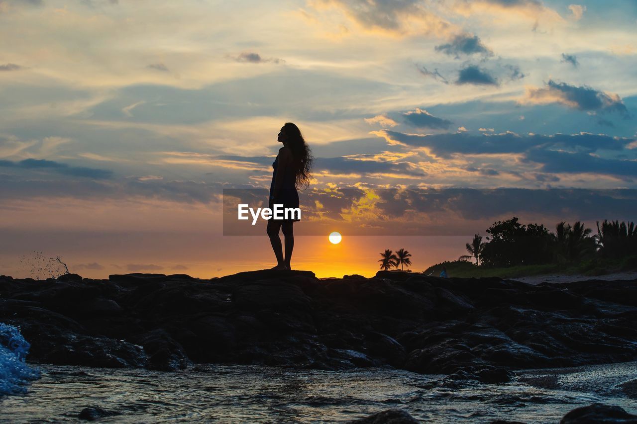 MAN STANDING ON ROCK BY SEA AGAINST SKY AT SUNSET