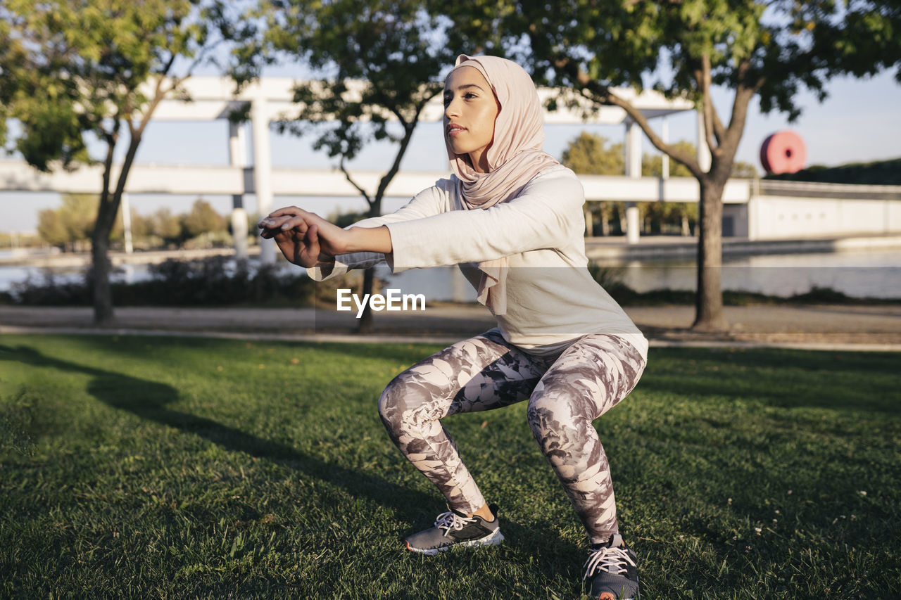 Full body of determined muslim female in hijab doing squat extending arms forward for balance during outdoor training