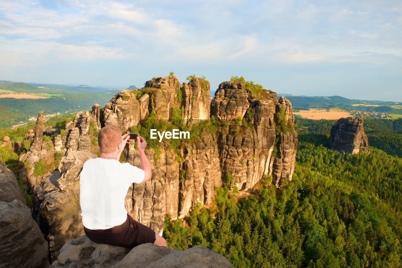 Short hair man on cliff of rock and takes photo by smart phone of land bellow. sunny day in rockd