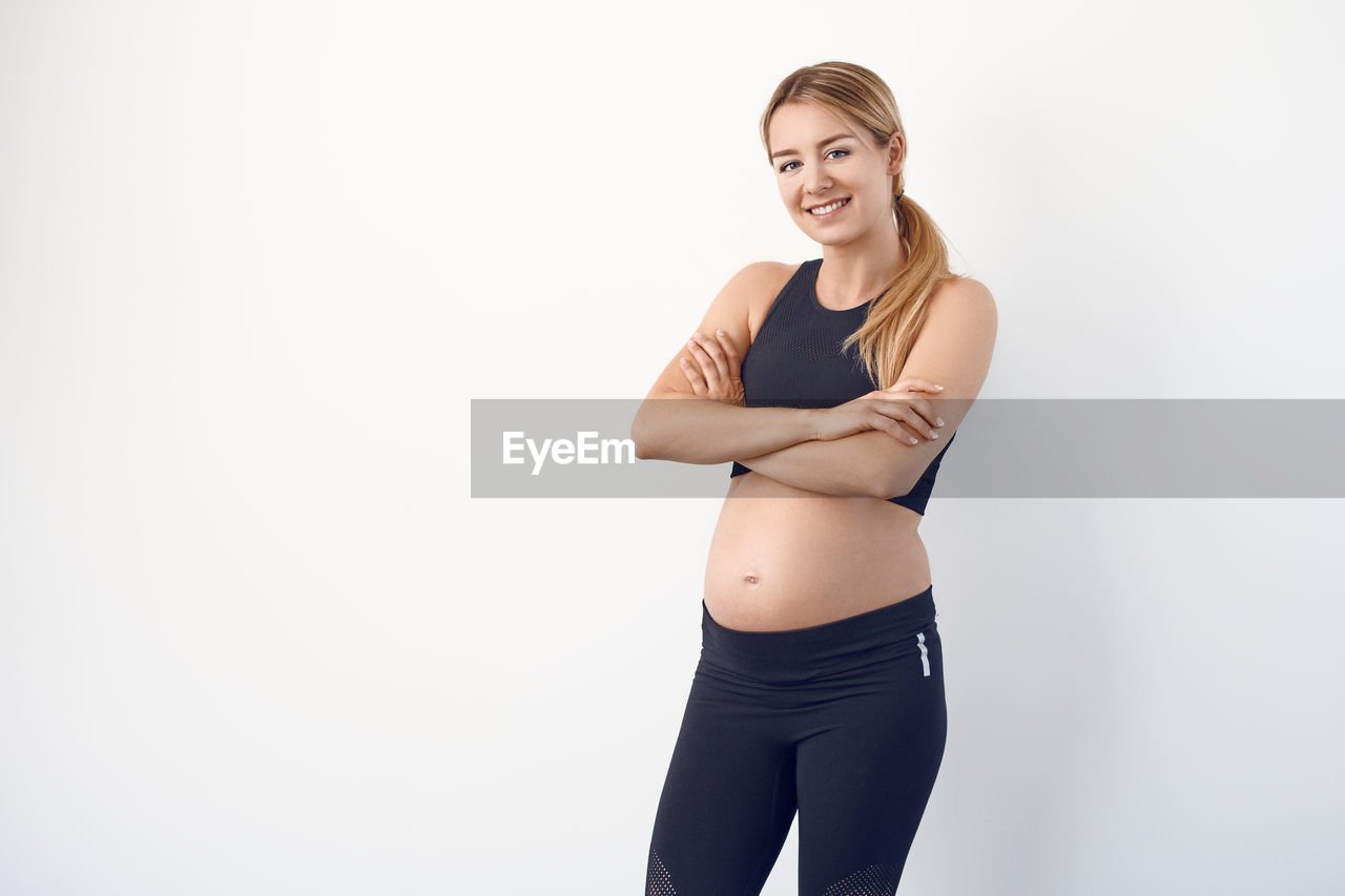 Portrait of smiling pregnant woman with arms crossed standing against white background