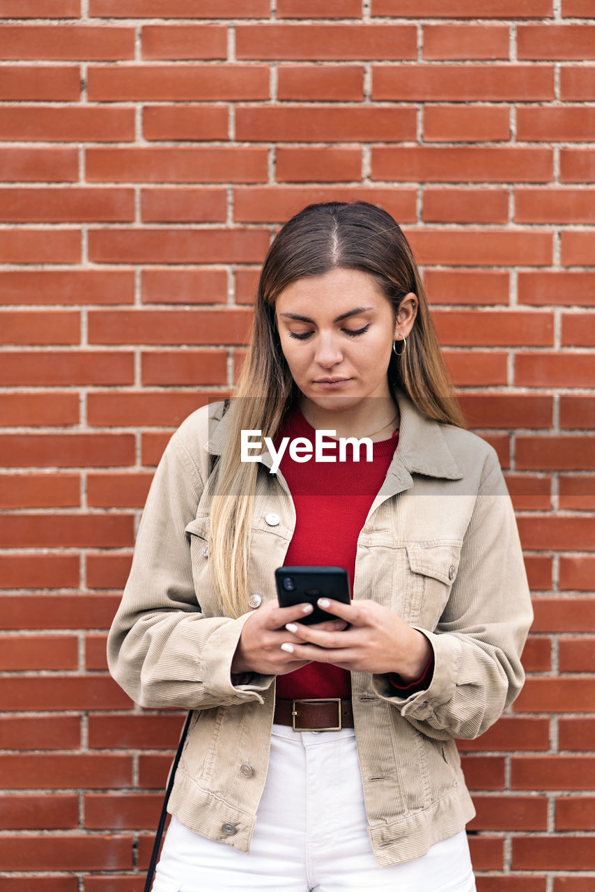 FULL LENGTH OF A YOUNG MAN USING PHONE AGAINST BRICK WALL