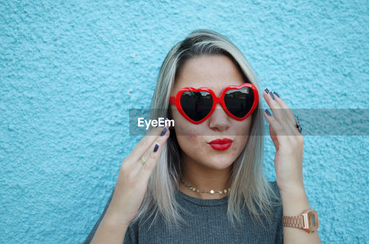 Young woman wearing heart shape sunglasses against blue wall