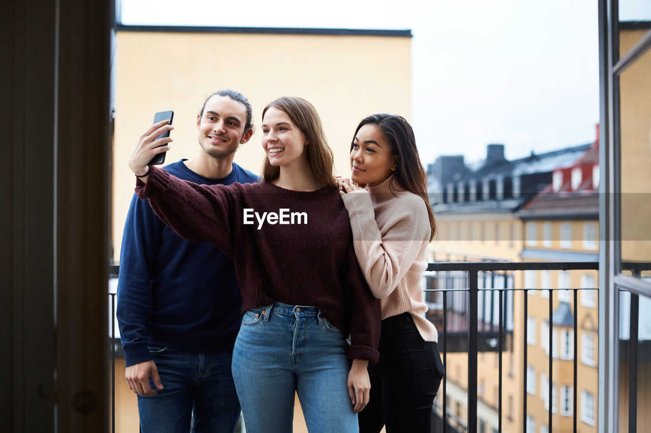 Smiling woman taking selfie with young friends while standing in balcony at rental apartment