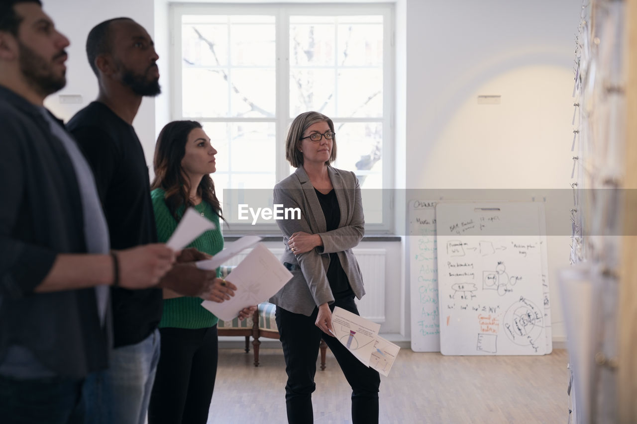 Multi-ethnic male and female engineers holding documents while reading notes on wall in office