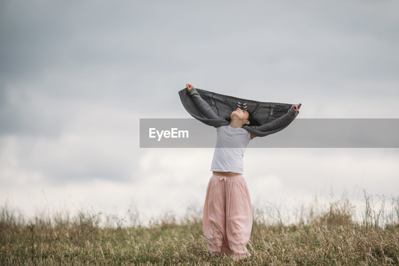 Girl holding sweater while standing on field against cloudy sky at park